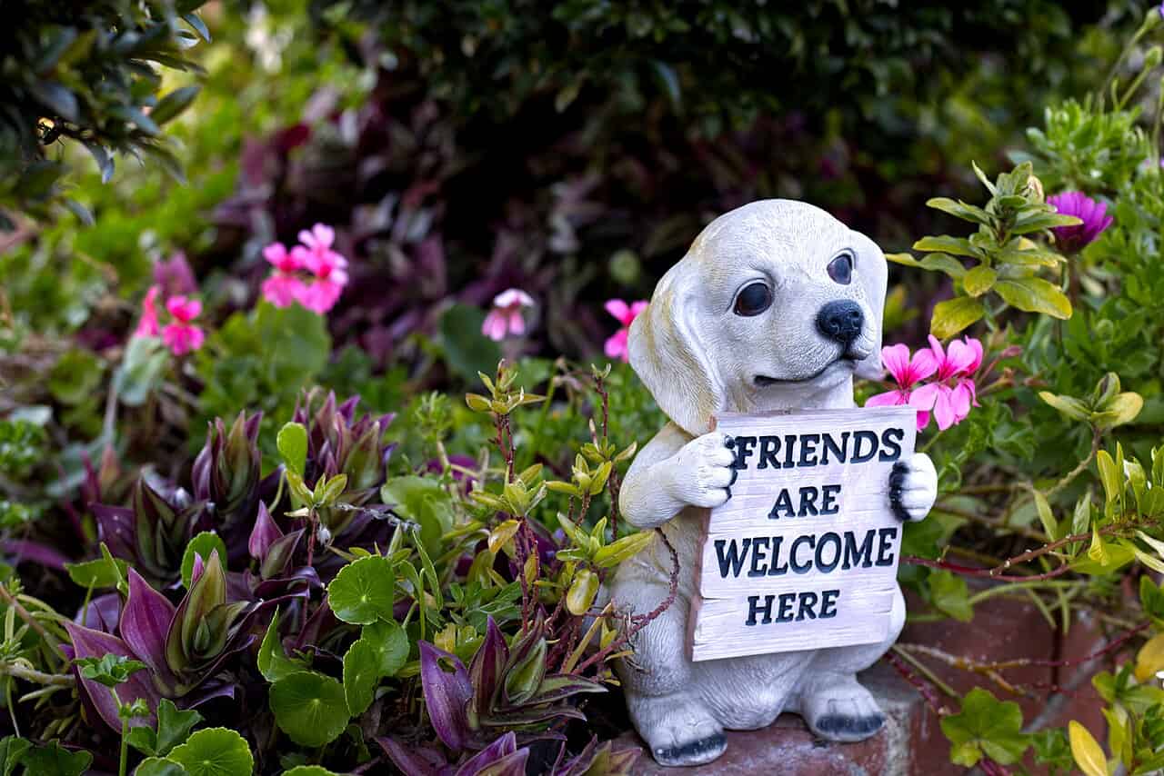 A small ceramic dog figurine holding a sign that reads "FRIENDS ARE WELCOME HERE," surrounded by vibrant green plants and pink flowers in a garden setting