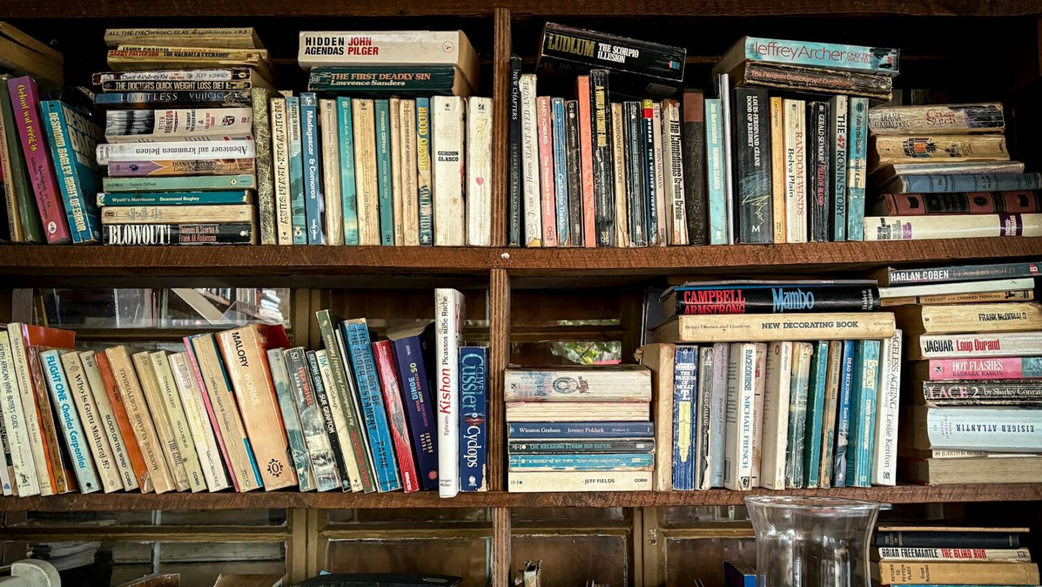A bookshelf filled with a variety of books, some stacked upright, others leaning slightly, titles visible on the spines, a mixture of genres, an older collection with a rustic wooden frame, a clear glass cup in the bottom corner, creating a cozy reading space