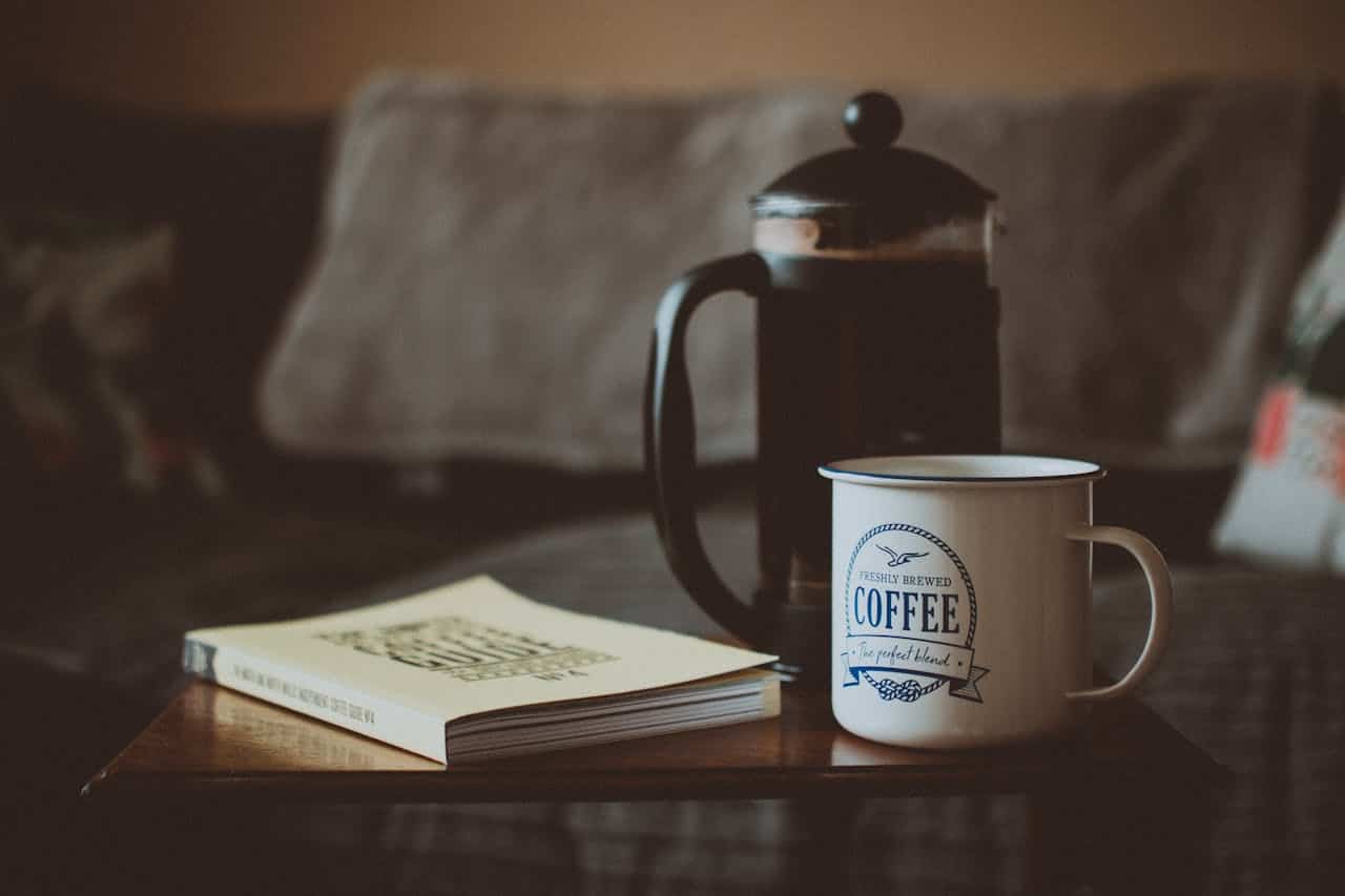 A cozy indoor scene featuring a French press, a white coffee mug labeled "Freshly Brewed Coffee," and a closed book on a wooden table, with a soft-focus sofa and cushions in the background