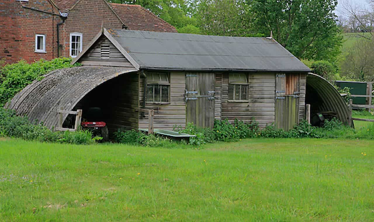 Weathered garden sheds with curved metal extensions on sides, situated on grassy lawn near brick farmhouse