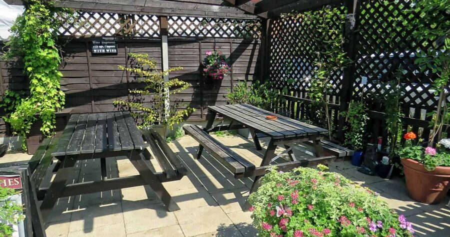 Outdoor seating area with wooden picnic tables under a partially covered pergola, lattice walls lined with green vines and potted flowering plants, designed as a cozy garden space for relaxation