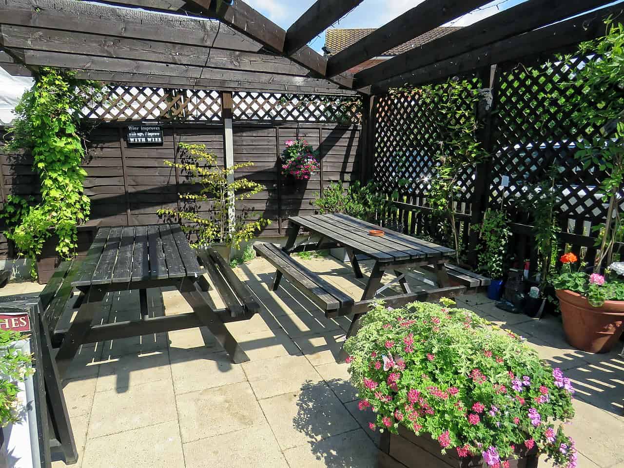 Outdoor seating area with wooden picnic tables under a partially covered pergola, lattice walls lined with green vines and potted flowering plants, designed as a cozy garden space for relaxation