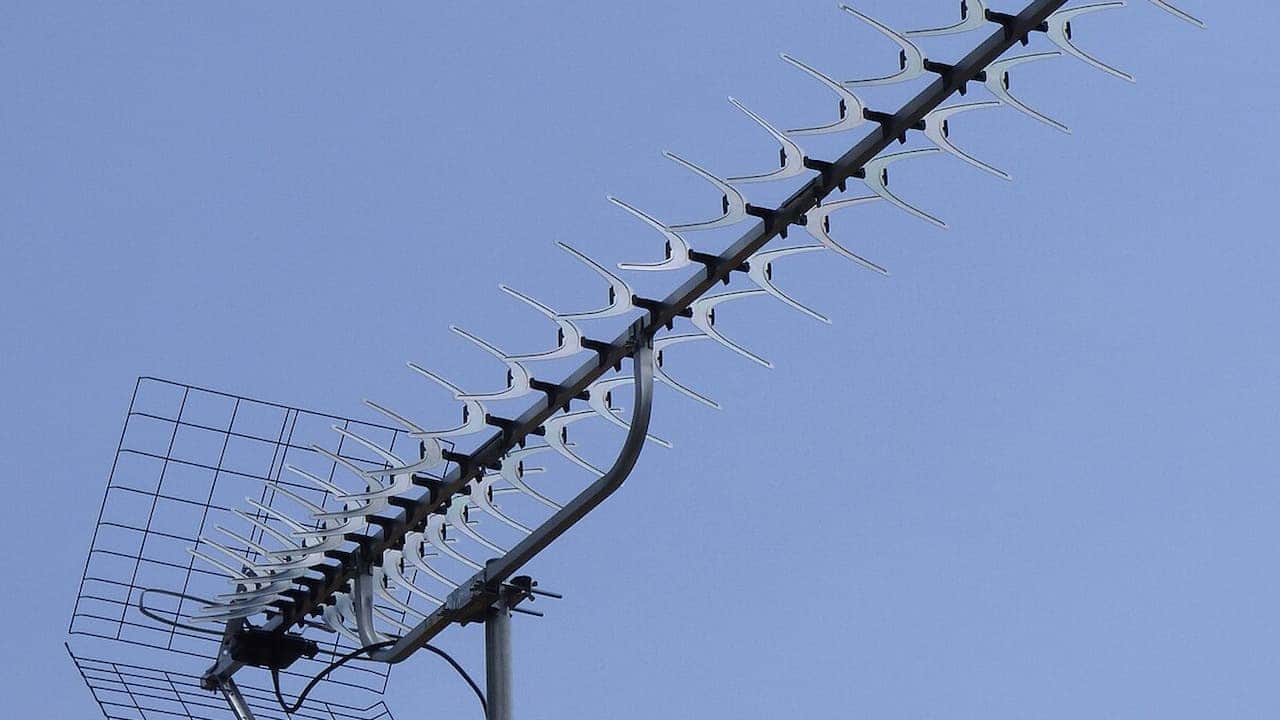 A high-definition rooftop TV antenna with multiple silver elements and a mesh reflector is mounted on a vertical pole against a clear blue sky, designed for optimal signal reception