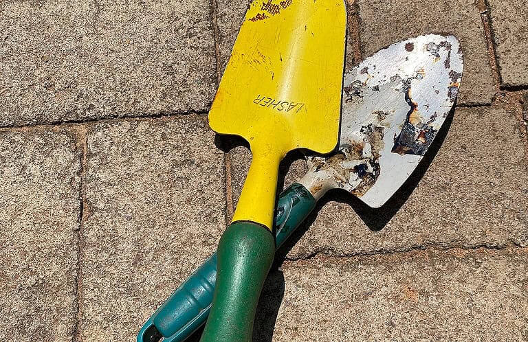 Two hand trowels with yellow and green handles, one with a worn, rusty metal surface and the other cleaner, lying on a paved surface, tools for gardening or digging