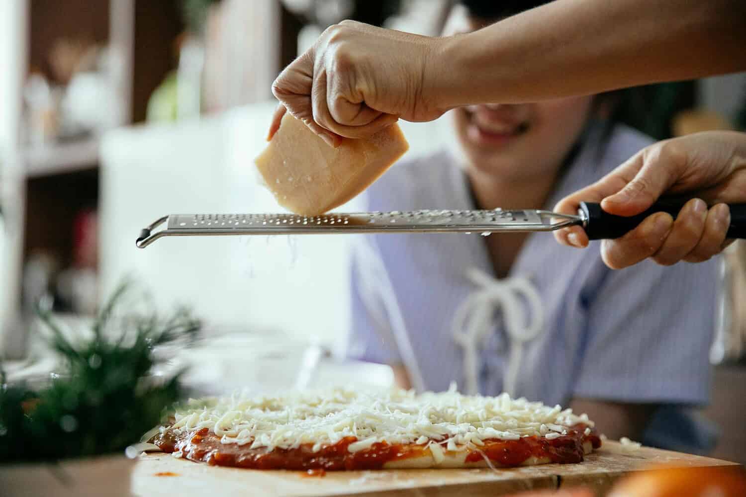 Hands grating cheese over a homemade pizza with a smiling person in the background.