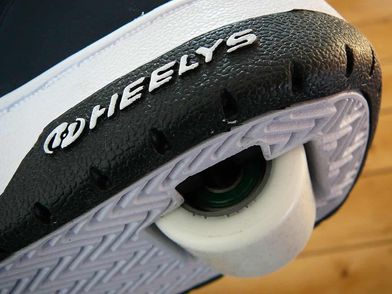 A close-up of a Heelys sneaker, showing the black and white sole with a visible wheel in the heel, the word "HEELYS" clearly displayed on the side, and the textured tread pattern on the bottom of the shoe