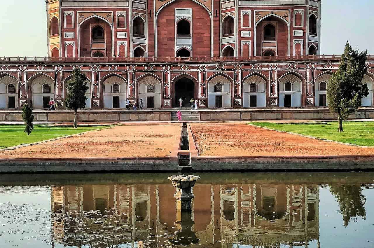 A view of Humayun's Tomb in Delhi, with its red sandstone architecture reflected in a water channel, people walking near the entrance, a stone pathway leading to the tomb, trees lining the garden, and a fountain in the foreground, showcasing the historical monument and its surroundings