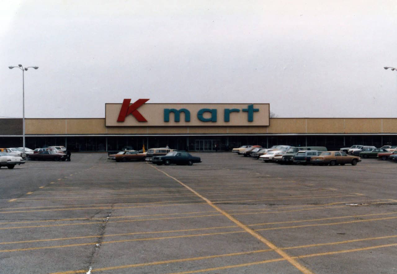 A Kmart store with large red and blue signage, a spacious parking lot filled with parked cars, a few people near the entrance, and a cloudy sky in the background, the store's exterior featuring a simple, retro design typical of older retail buildings