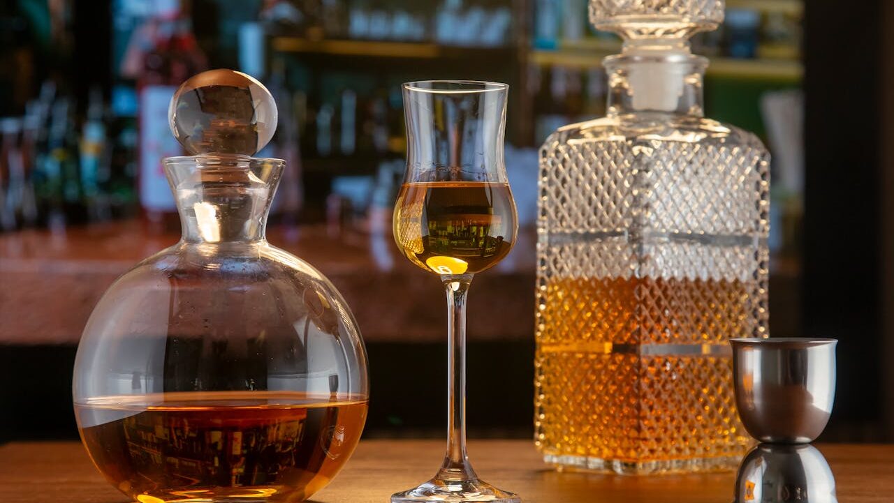 A wooden table with three elegant glass containers—two decanters and one tall liqueur glass—filled with amber liquor, alongside a metal jigger. The blurred background displays a bar shelf stocked with various bottles