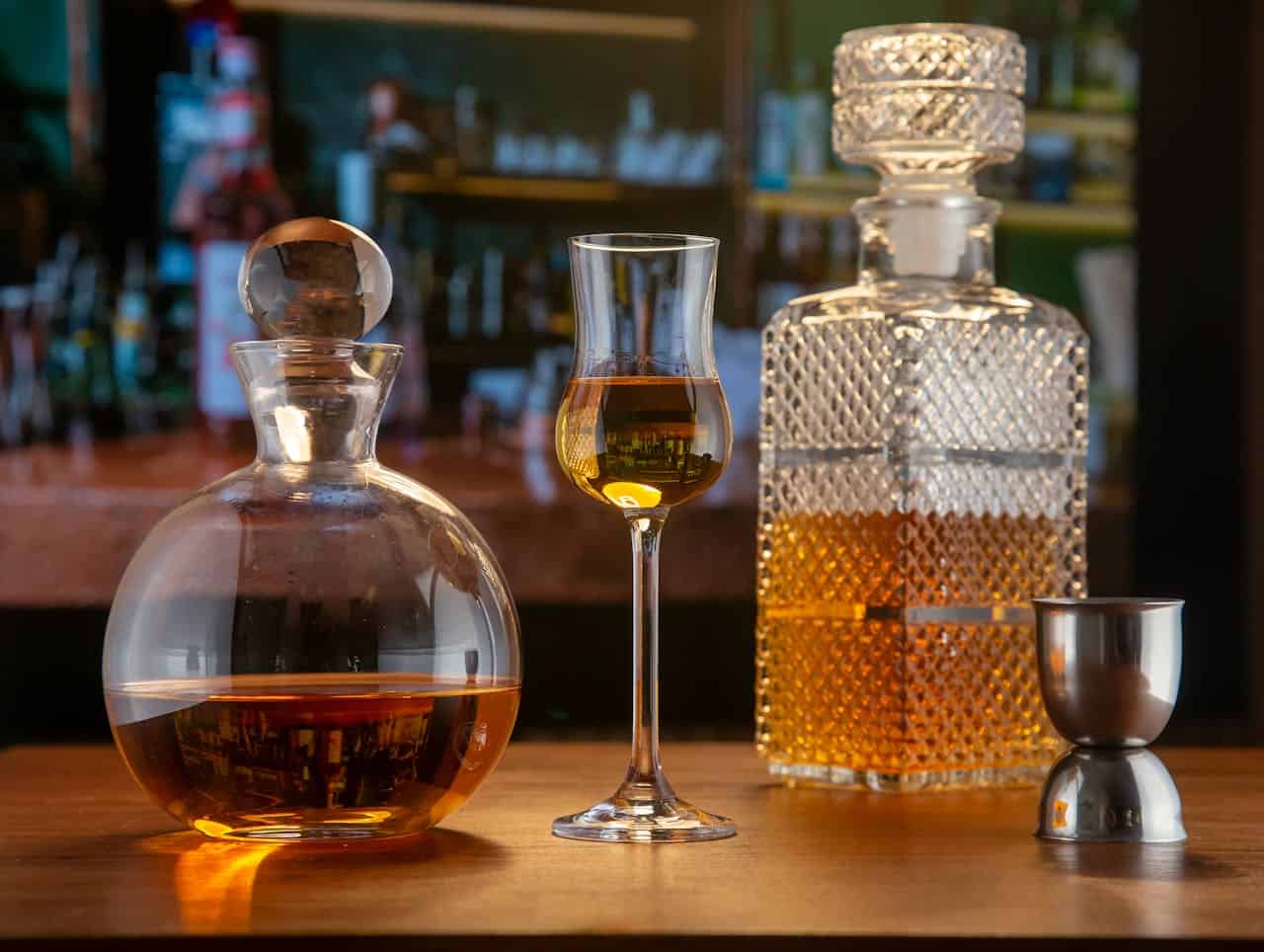 A wooden table with three elegant glass containers—two decanters and one tall liqueur glass—filled with amber liquor, alongside a metal jigger. The blurred background displays a bar shelf stocked with various bottles