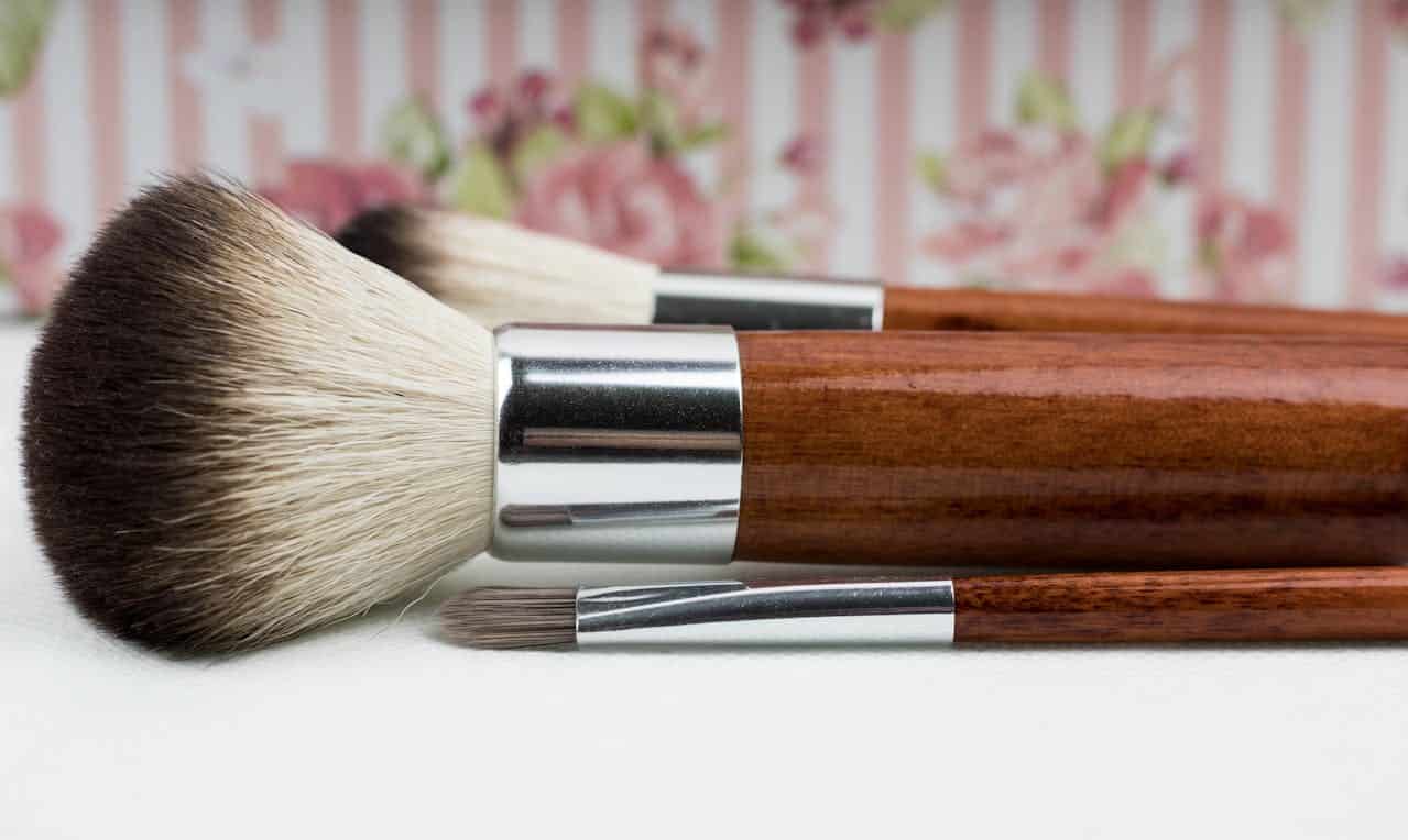 Makeup brushes with wooden handles and silver ferrules, featuring different sizes and bristle types, arranged against a floral pink striped background