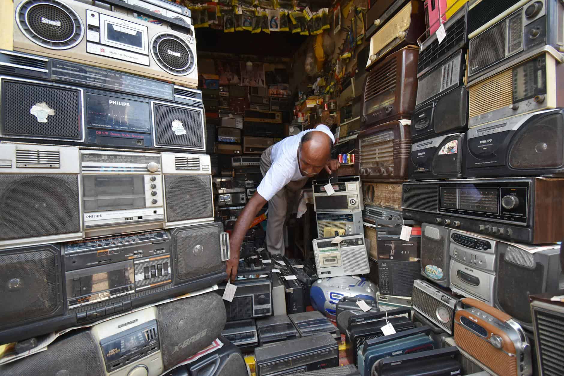Man in Kolkata shop filled with vintage radios and electronics. Nostalgic atmosphere.