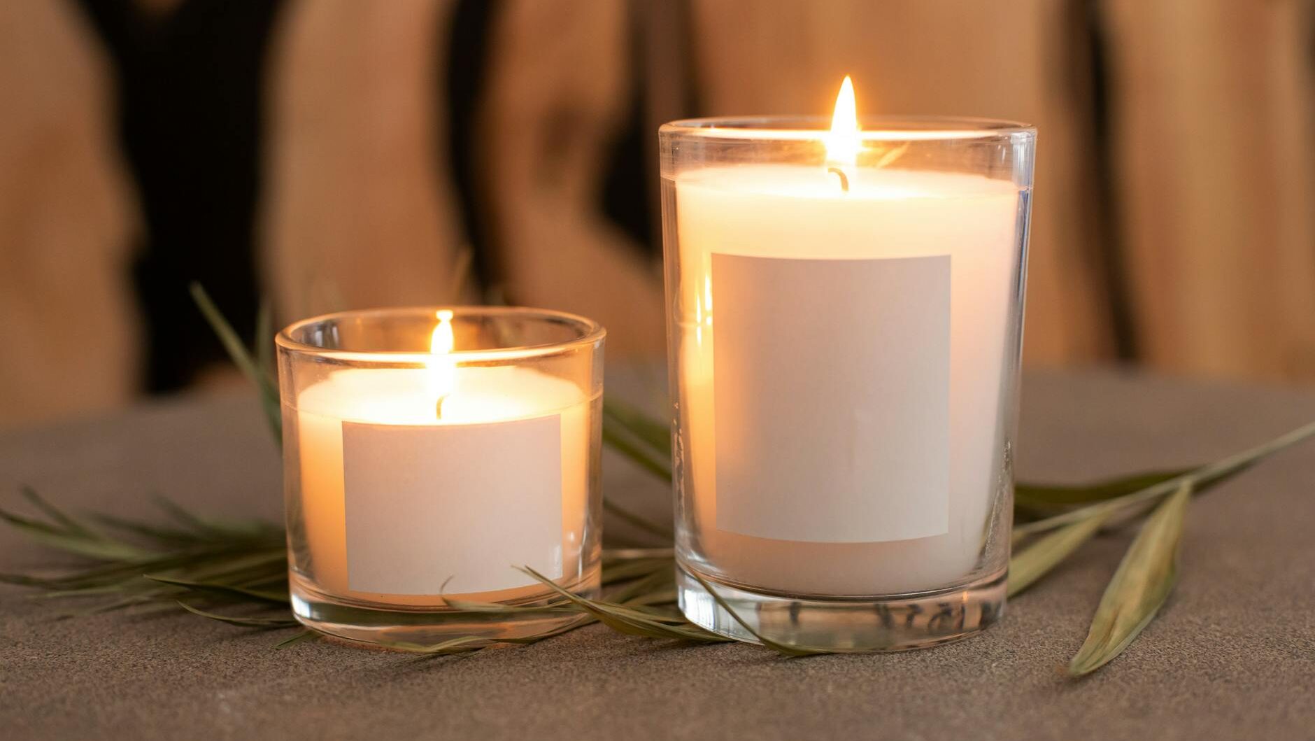 Minimalist candles in glass containers with blank labels, surrounded by leaves.
