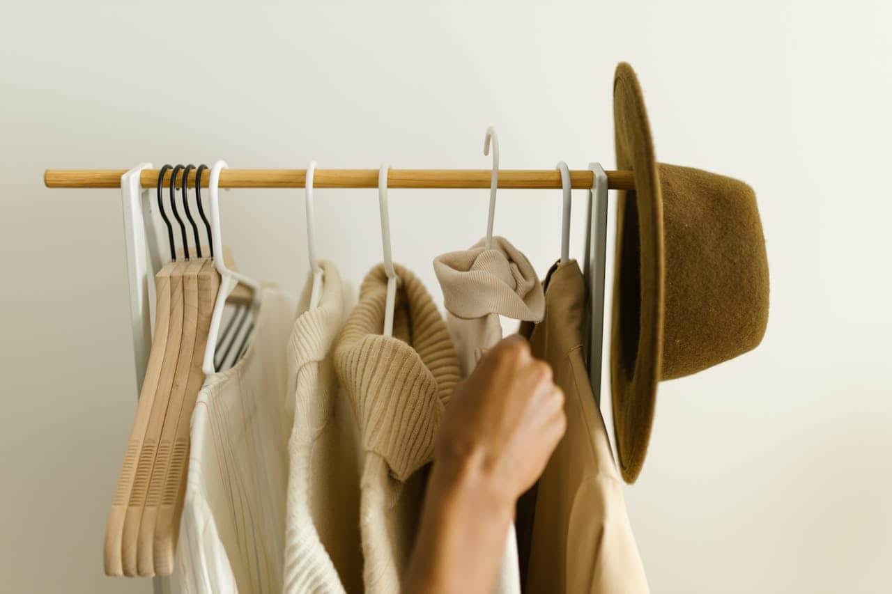 A minimalist wooden clothing rack with neutral-toned garments on white and black hangers. Cream sweaters and tops are visible alongside a brown felt hat. A hand reaches to touch an item