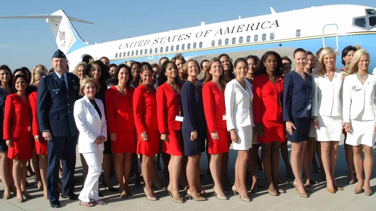 A group of women, wearing red, navy, and white suits, posing together in front of a U.S. Air Force plane, some smiling and standing in a row, others clapping and cheering, with a bright, clear sky in the background, showcasing a formal event or photoshoot