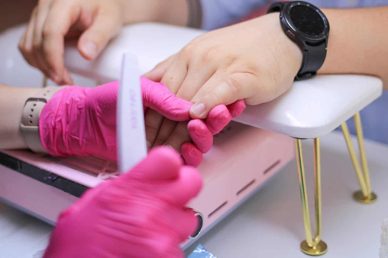 Closeup of nail technician wearing bright pink gloves filing client's nails, client's hand resting on white manicure armrest with gold legs, technician and client both wearing watches