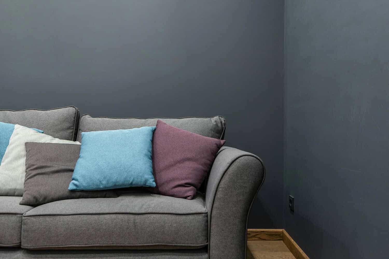 Part of soft modern gray sofa with armrest and abundance of bright pillows placed near wall in light room at home