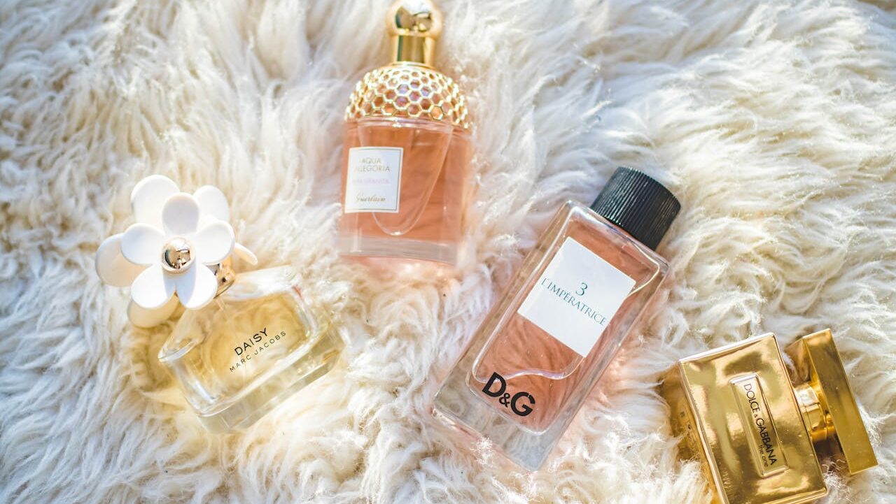 Four luxury perfume bottles arranged on white fluffy rug, including Marc Jacobs Daisy with flower cap, Guerlain Aqua Allegoria, D&G L'Imperatrice, and Dolce & Gabbana gold bottle, elegant fragrance display