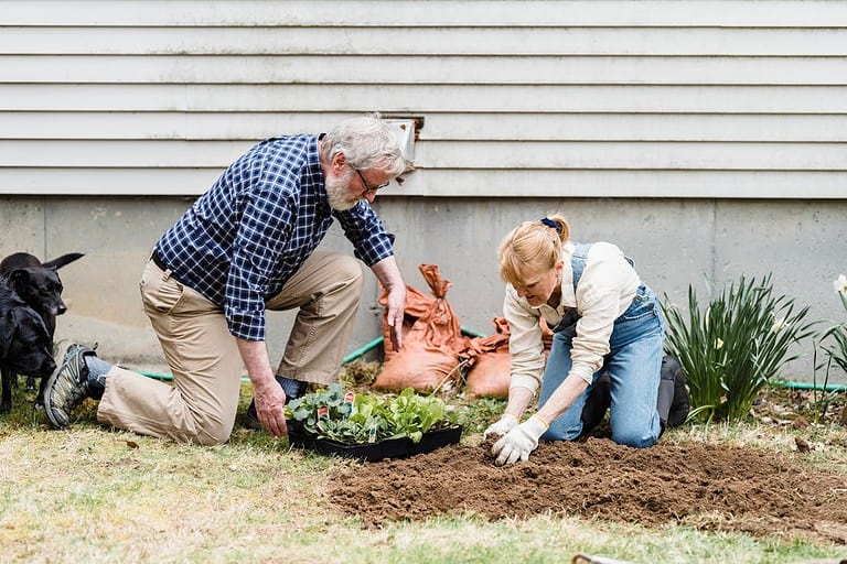 Older man in plaid shirt and younger woman with blonde hair gardening together, both kneeling beside prepared soil, seedling tray nearby, black dog watching, house siding in background