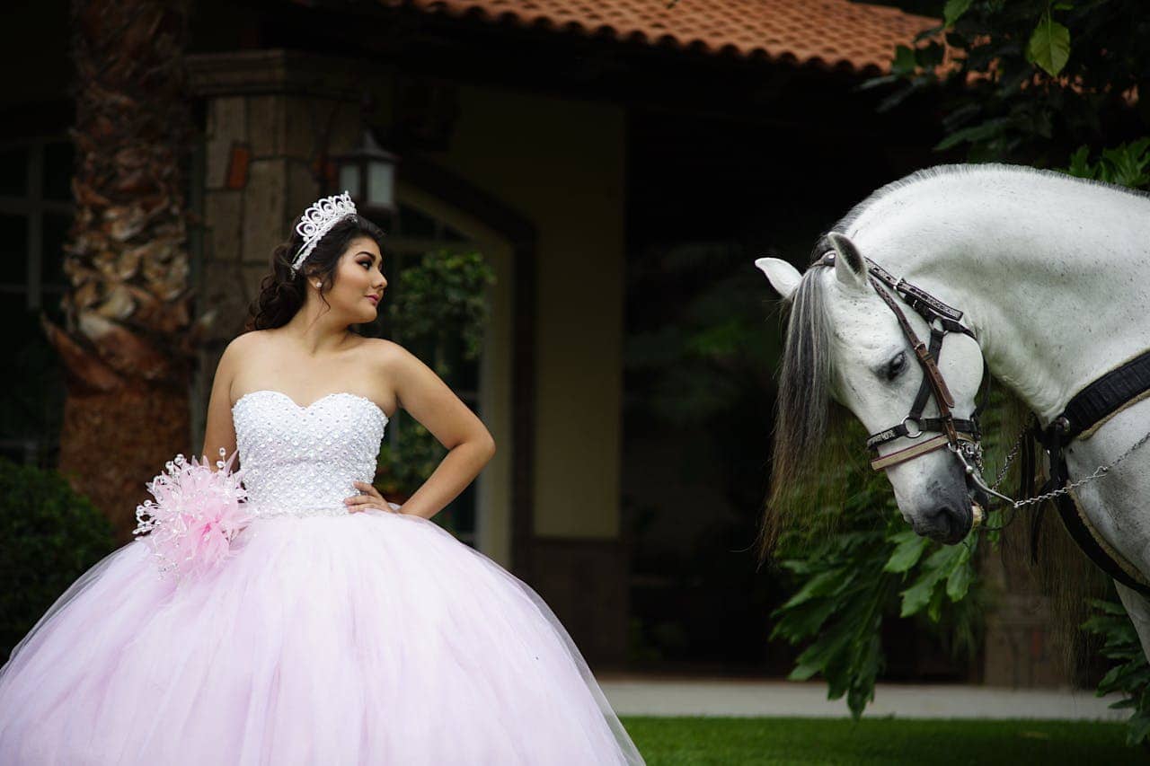 Young woman in pink quinceañera dress with tiara and bouquet standing beside white horse, Spanish-style building with terra cotta roof in background, formal celebration photoshoot
