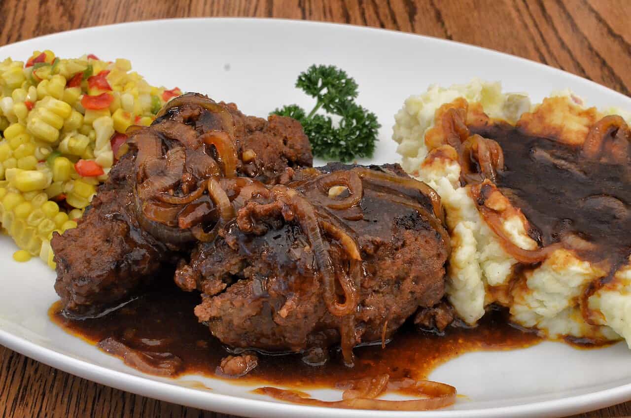 A plate with Salisbury steak covered in onions and gravy, served with mashed potatoes and a side of colorful corn salad, garnished with a parsley sprig
