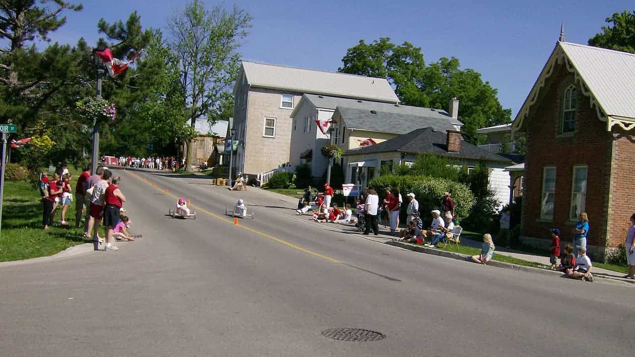 A street lined with spectators watching a soapbox derby, children racing small, homemade vehicles down the road, people sitting along the sidewalk in the sun,
