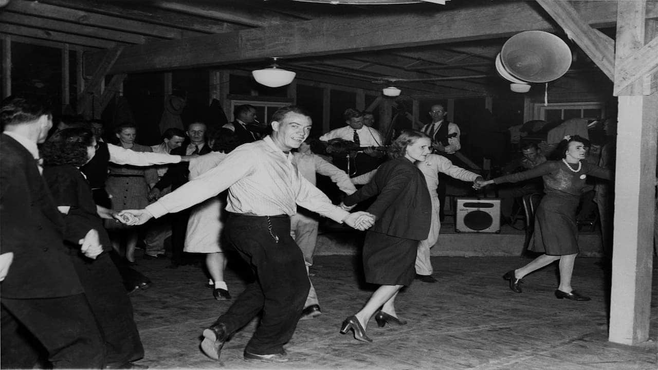 A group of people square dancing in a barn-like hall, holding hands and performing synchronized movements, musicians playing instruments in the background