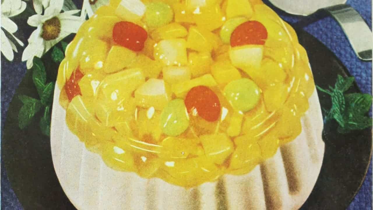 A close-up of a colorful fruit gelatin dessert on a plate, featuring yellow gelatin with chunks of fruit like grapes and cherries, served on a white dish, decorated with a small flower arrangement in the background, vintage styling