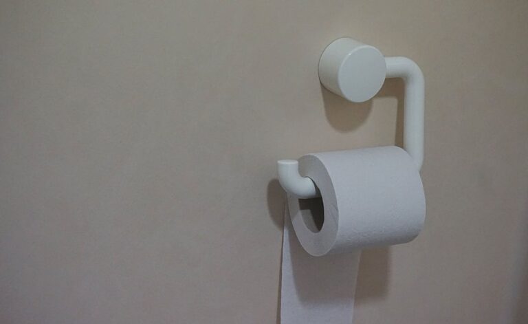 A minimalist white toilet paper holder mounted on a beige wall. The holder has a simple, modern design with a circular wall mount and curved arm supporting a roll of white toilet paper