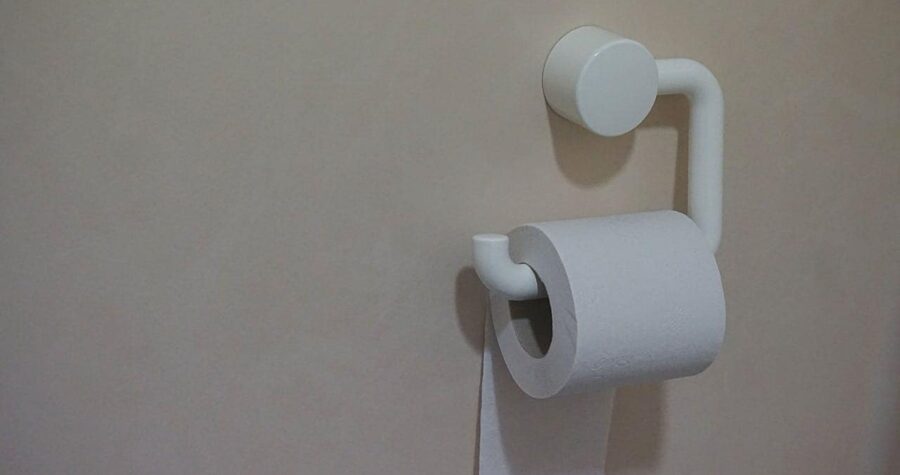 A minimalist white toilet paper holder mounted on a beige wall. The holder has a simple, modern design with a circular wall mount and curved arm supporting a roll of white toilet paper