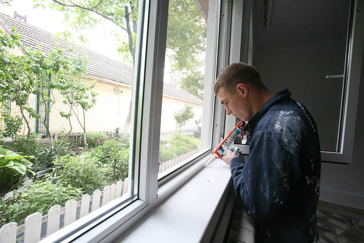 A man in a blue work jacket, applying sealant with a caulking gun along a window frame, focused on the task, a white picket fence and green garden visible outside the window, with a bright, natural setting