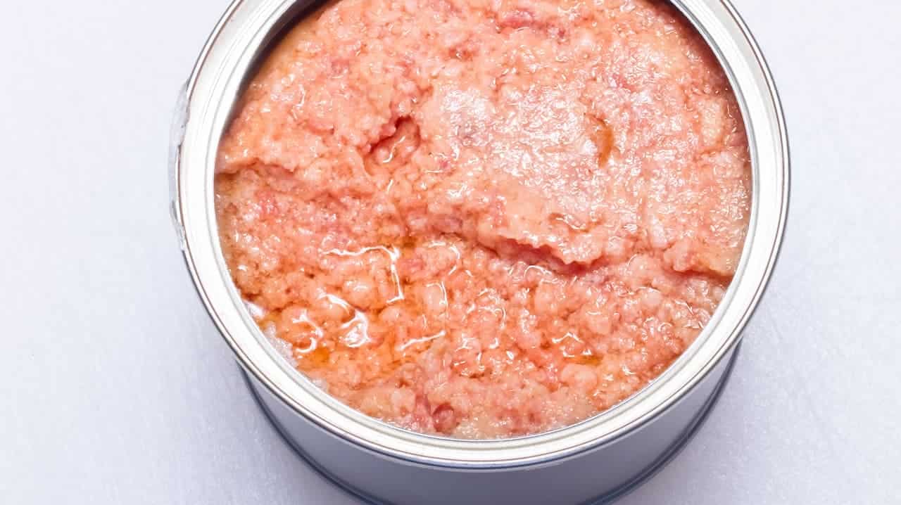 Deviled ham in a metal can, finely mashed texture, visible oily sheen, light pink color with slight moisture, processed meat spread, ready for use in sandwiches or dips, compacted texture with a smooth consistency, food product in a sealed container, classic preserved snack