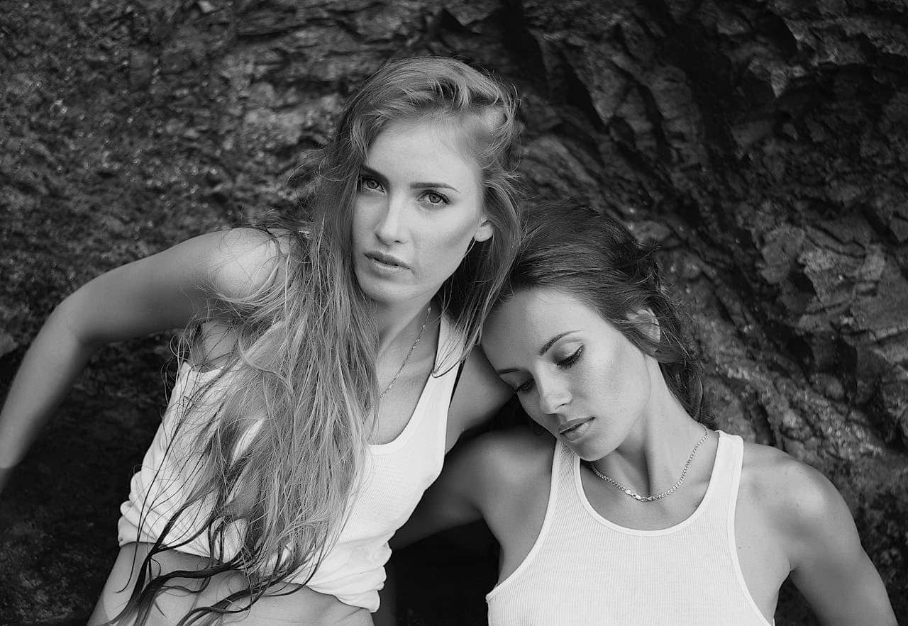 Black and white portrait of two women in white tank tops against a rocky backdrop. One has long hair looking at camera, while the other has eyes closed resting against her companion's shoulder
