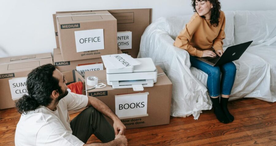 Man and woman sitting on the floor among labeled moving boxes in a room with wooden floors, surrounded by packed items including a printer and tape, appearing to take a break or chat during the process of moving or unpacking