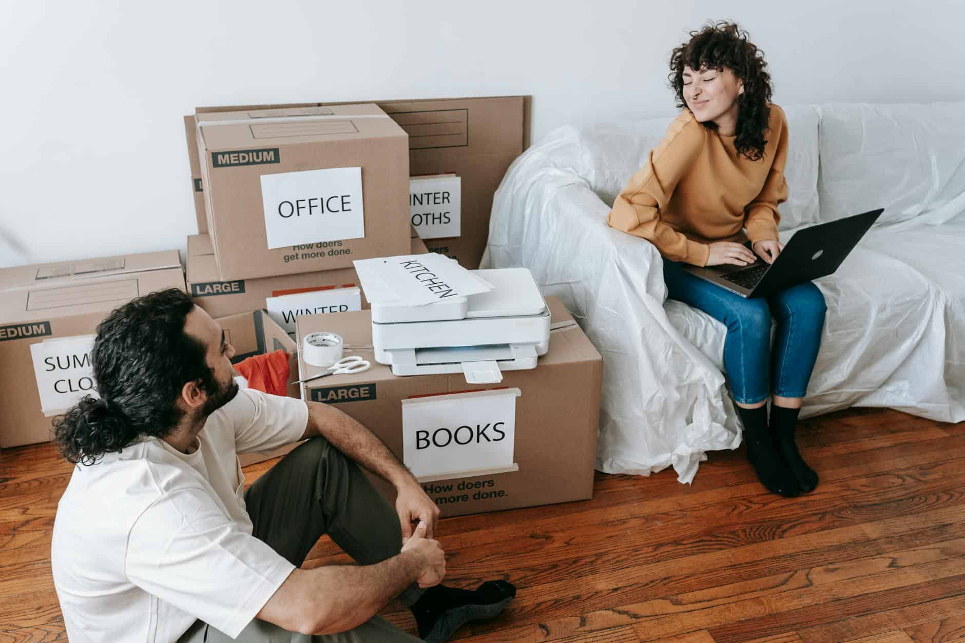 Man and woman sitting on the floor among labeled moving boxes in a room with wooden floors, surrounded by packed items including a printer and tape, appearing to take a break or chat during the process of moving or unpacking