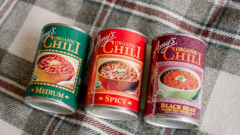 Three Amy’s Organic Chili cans—Medium, Spicy, and Black Bean—sit on a plaid fabric surface, slightly tilted forward