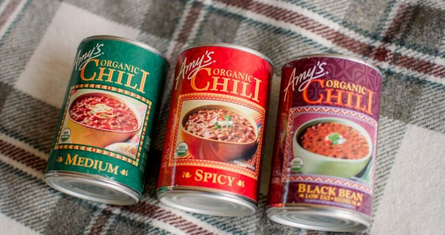 Three Amy’s Organic Chili cans—Medium, Spicy, and Black Bean—sit on a plaid fabric surface, slightly tilted forward