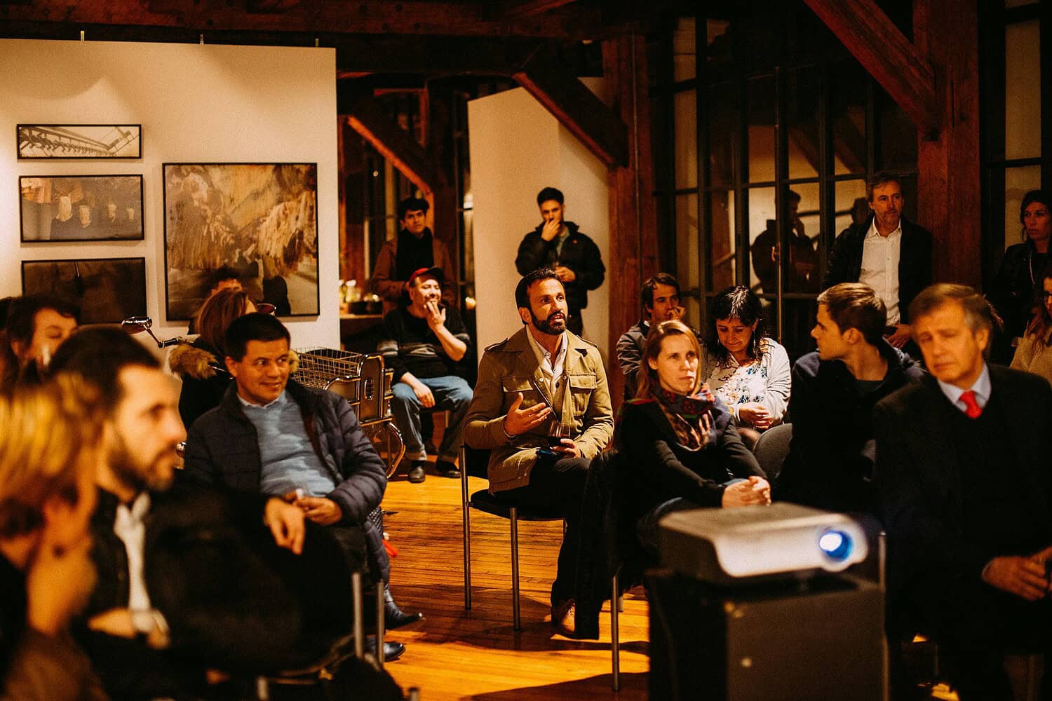 A group of people attending a townhall meeting in a well-lit space, seated in chairs, some participants engaged in conversation while others are listening, the setting features art on the walls and a projector in the foreground displaying content to the audience, a casual and relaxed atmosphere