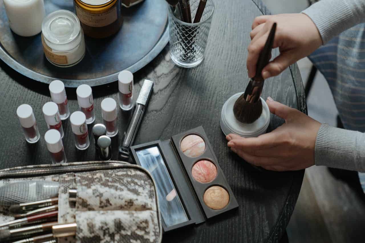 Person in gray sweater using makeup brush with loose powder, surrounded by cosmetics including eyeshadow palette, lipstick tubes, makeup bag with brushes, on dark wooden surface