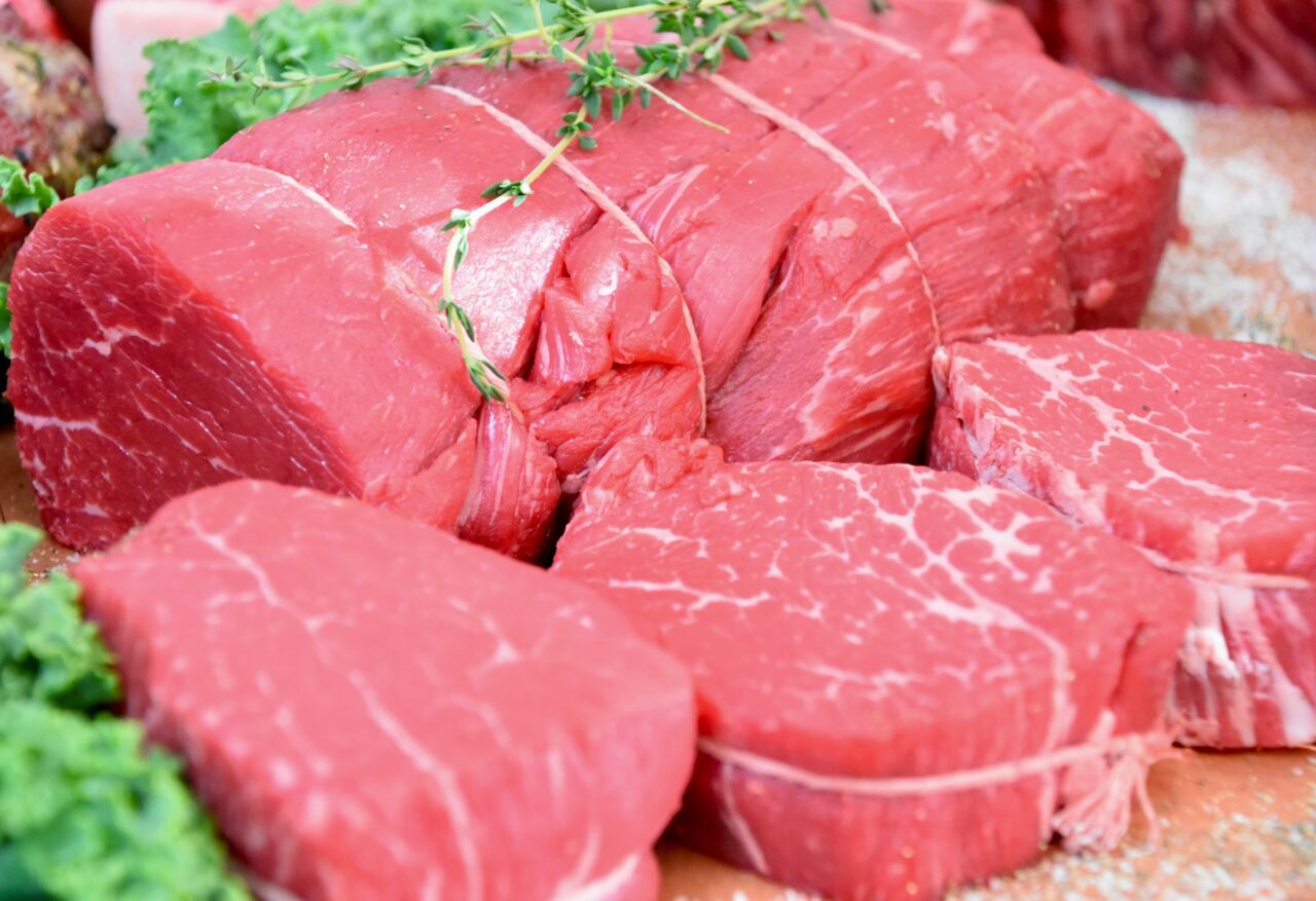 The image shows cuts of raw beef steak with visible marbling, a sprig of thyme is tied to one of the larger cuts, the steaks are neatly arranged, with the background blurred, some leafy greens are also visible, providing a fresh and clean presentation of meat for culinary purposes