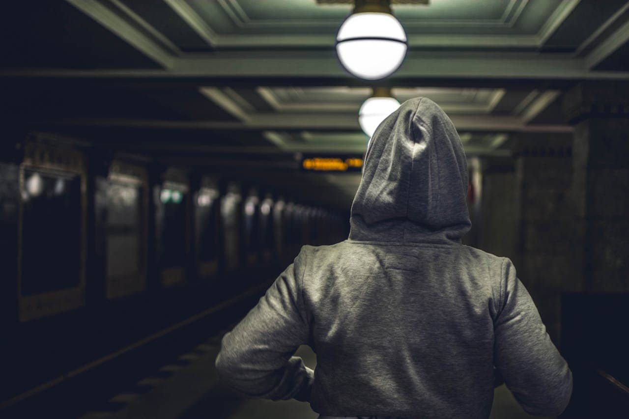 Person in gray hoodie viewed from behind in dimly lit subway station, globe lights on ceiling, empty platform, digital display sign visible in distance, moody atmosphere