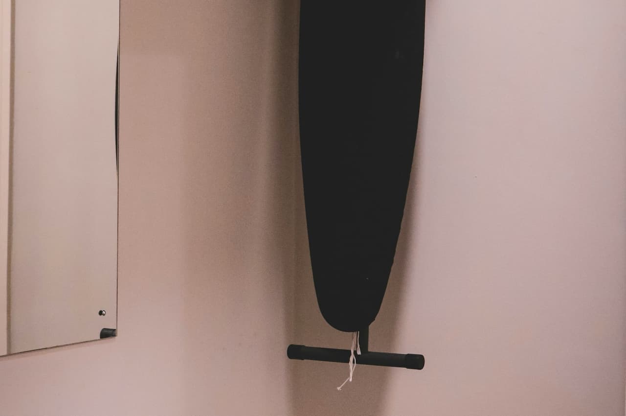 The image depicts a small closet area with an iron board hanging on the wall, a pair of black shoes lying on the floor, a mirror reflecting part of the scene, a stool, and a coat rack with empty hangers