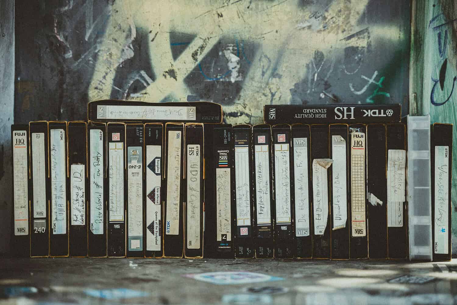 A collection of VHS tapes stacked neatly, each with handwritten labels on the spines, various brands like Fuji and TDK visible, some tapes are partially worn, with a background featuring graffiti, evoking a sense of nostalgia for older media storage