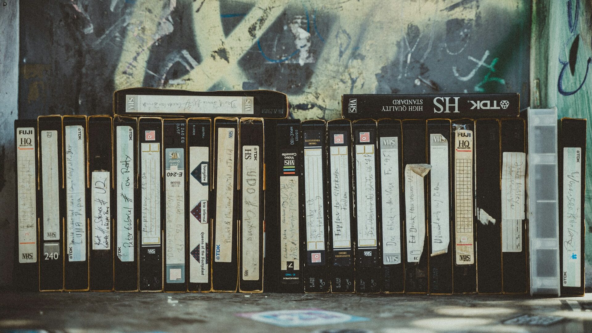 A collection of VHS tapes stacked neatly, each with handwritten labels on the spines, various brands like Fuji and TDK visible, some tapes are partially worn, with a background featuring graffiti, evoking a sense of nostalgia for older media storage