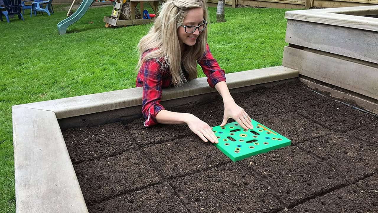 A woman in a red plaid shirt using a green garden tool with multiple holes to space out seeds in a raised garden bed, smiling while preparing the soil for planting, in a well-maintained garden with grass and a playground in the background