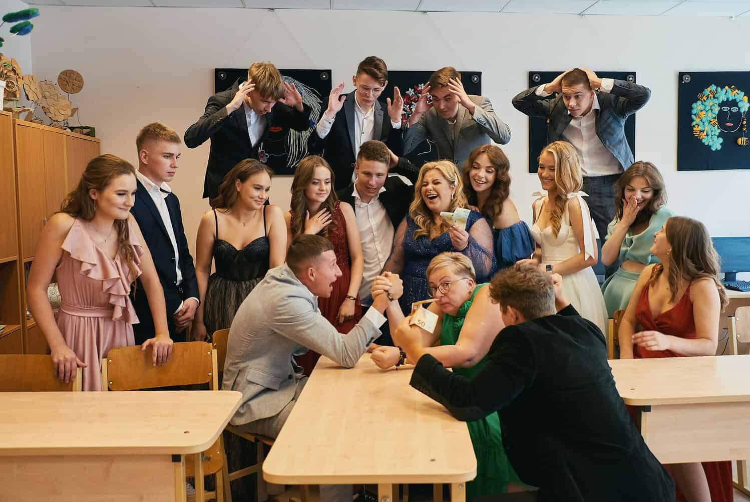 A group of people posing for a photo in a classroom, some standing and some seated, a friendly atmosphere, two people engaging in an arm-wrestling competition, others laughing and enjoying, one woman holding money, dressed in formal and party attire, vibrant and joyful scene