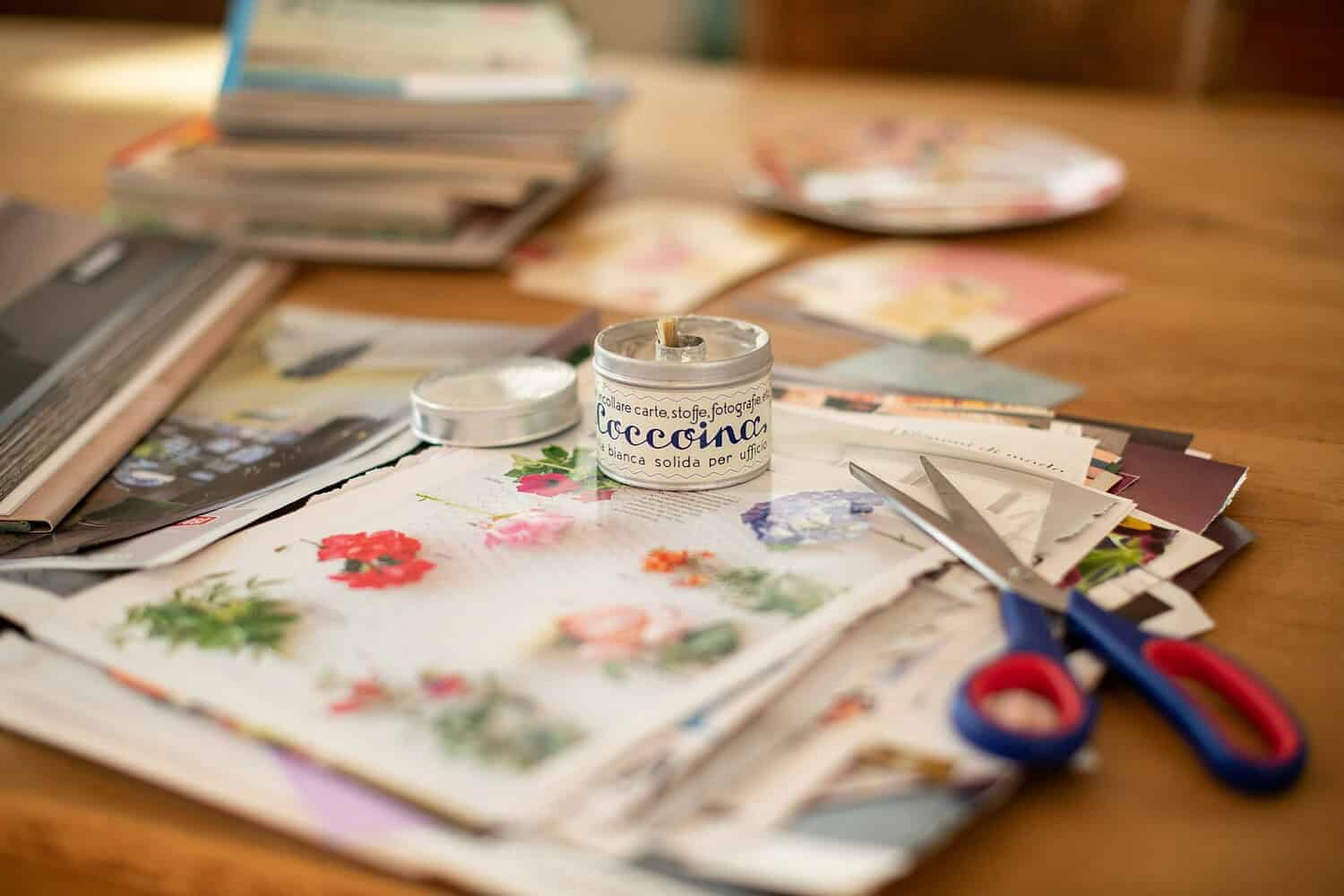 A collection of various magazines and paper scraps spread on a wooden table, a tin of white glue, scissors with red and blue handles, paper with floral prints, crafting materials ready for a creative project such as scrapbooking