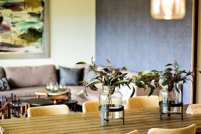 Modern dining area with wooden table, yellow chairs, glass vases containing eucalyptus leaves, beige sofa in background, abstract artwork on wall, contemporary interior design