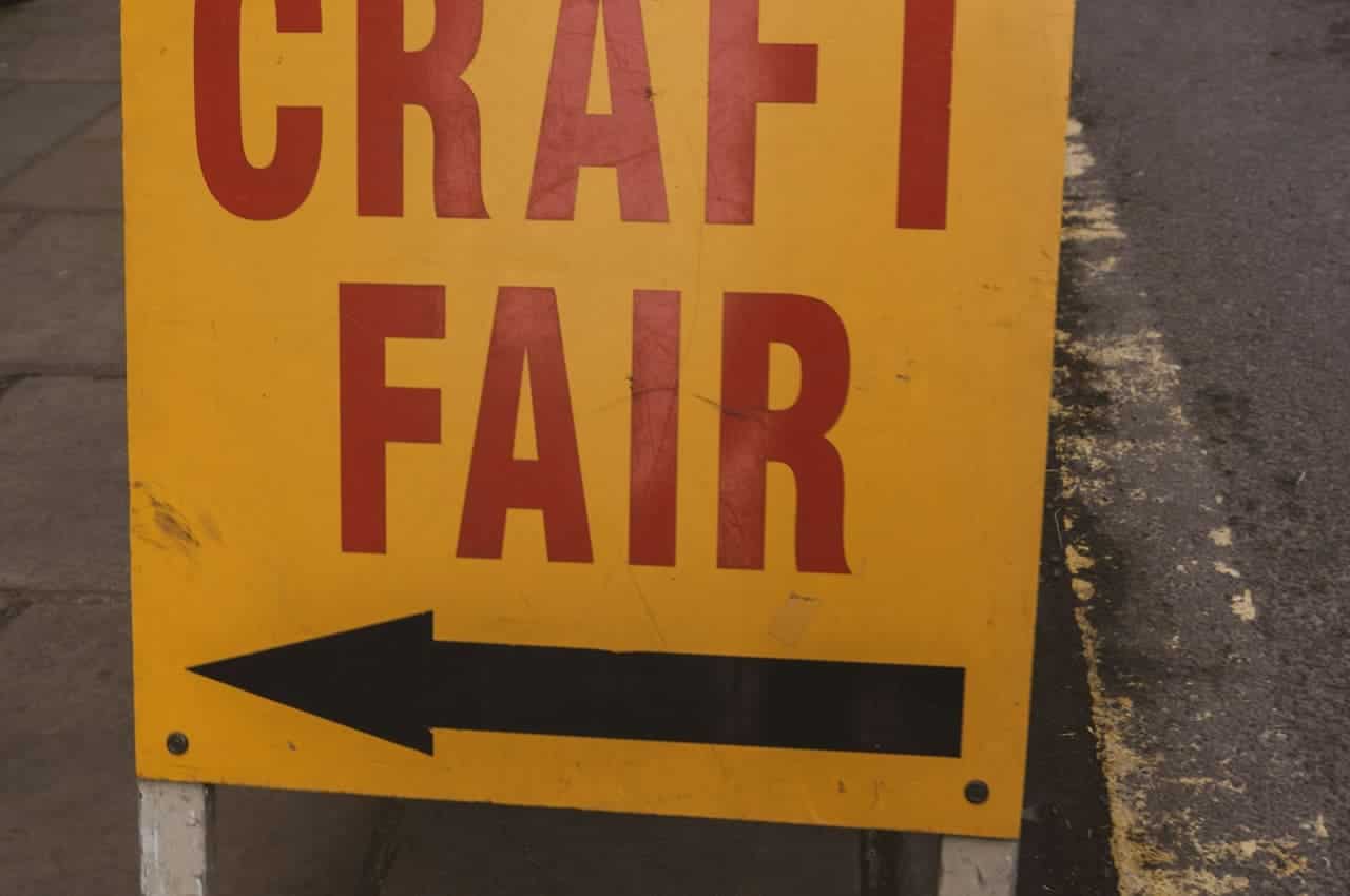 Yellow sign with bold red text saying "CRAFT FAIR," left-pointing black arrow, sign directing people to the location of a craft fair, placed on the sidewalk, clear and bright lighting, street and pavement visible in the background