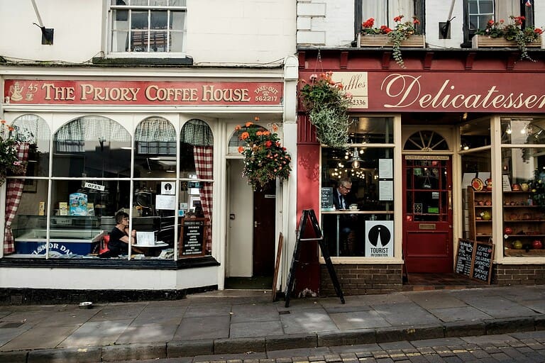 A vibrant street scene showing two shops, "The Priory Coffee House" on the left with a woman sitting by the window and "Delicatessen" on the right with a man enjoying a beverage, both shops have plants hanging above the entrances, a tourist information sign, and a clear view of the cobbled street outside
